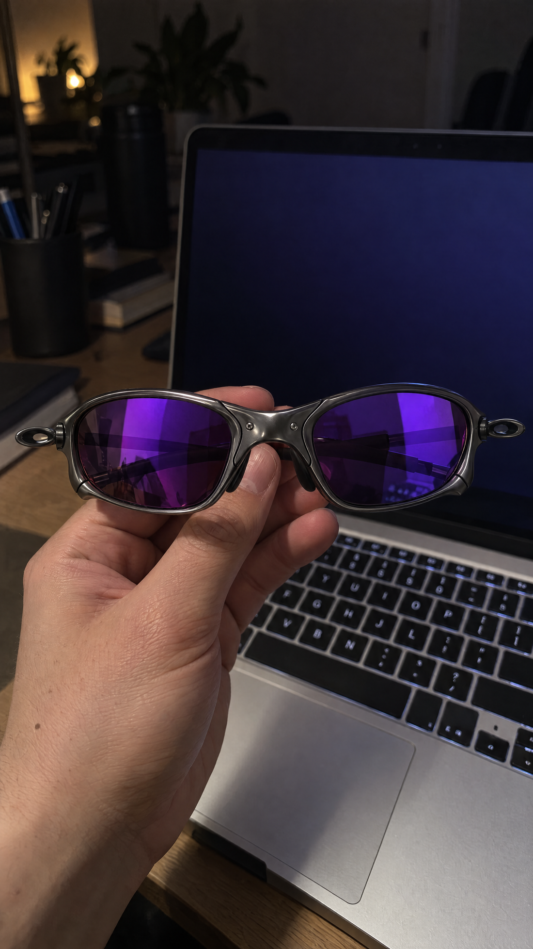 Hand holding Houser metal sunglasses with Purple Mirror lenses in front of a laptop