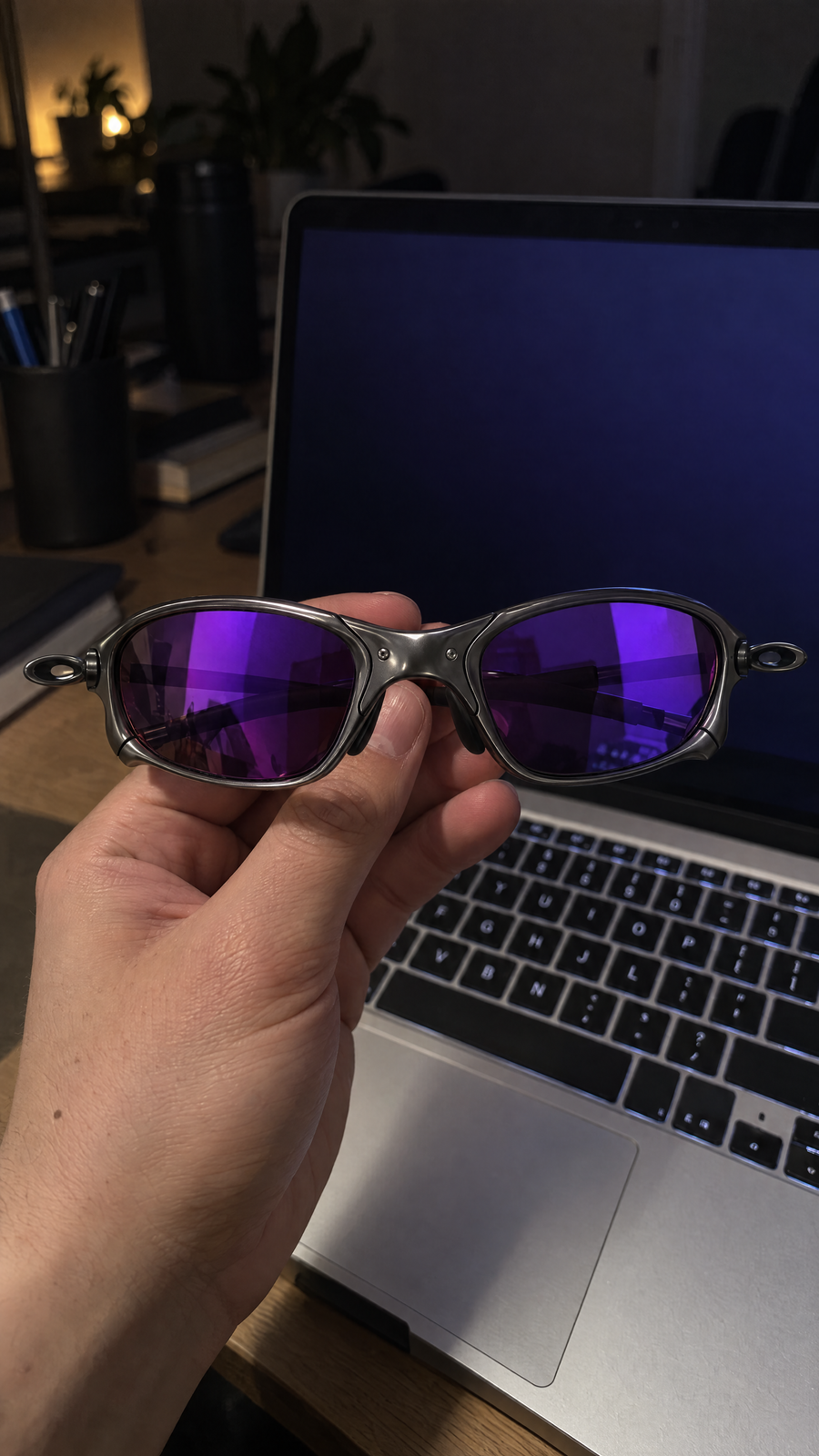 Hand holding Houser metal sunglasses with Purple Mirror lenses in front of a laptop